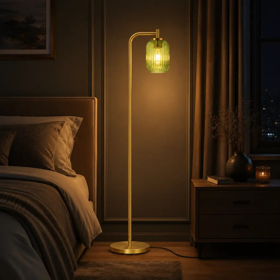 A modern floor lamp with a slim gold metal stand and a ribbed green glass shade glowing with warm light, placed in a dimly lit bedroom next to a bed and nightstand.