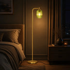 A modern floor lamp with a slim gold metal stand and a ribbed green glass shade glowing with warm light, placed in a dimly lit bedroom next to a bed and nightstand.