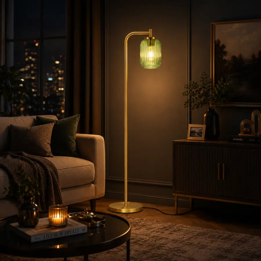 A modern floor lamp with a gold-finished metal frame and a ribbed green glass shade glowing with warm light, placed in a dimly lit living room next to a sofa and console table.