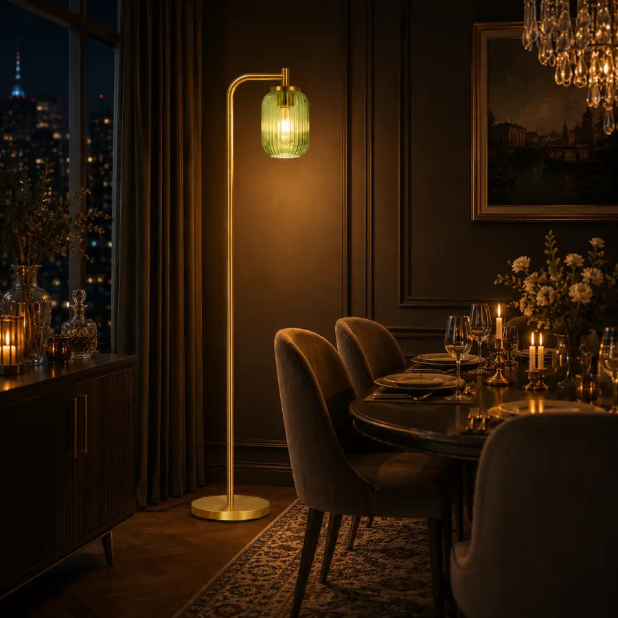 A modern floor lamp with a curved gold-toned metal stand and a ribbed translucent green glass shade glowing with warm light, placed in a dark, elegantly furnished dining room next to a window with a city view.
