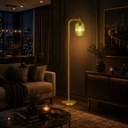 A modern floor lamp with a slim gold metal stand and a textured green glass shade glowing with warm light, placed in a stylishly decorated living room next to a sofa and console table.