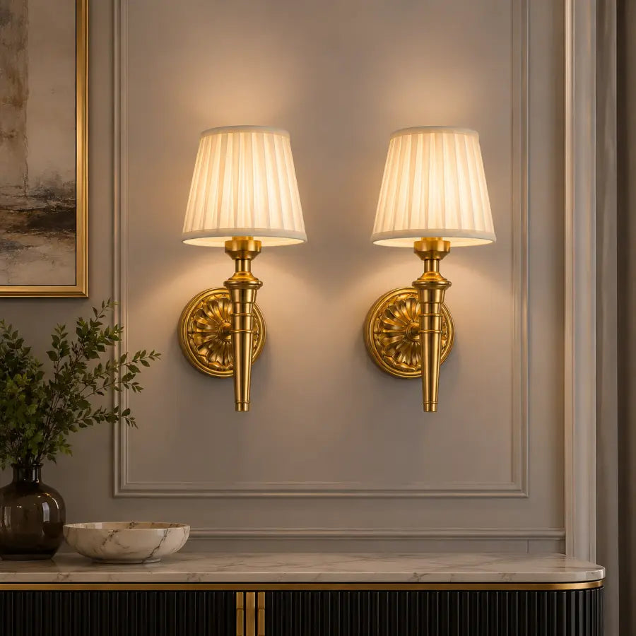 Two matching wall sconces with pleated white fabric shades and ornate gold metal bases mounted on a gray paneled wall above a marble console table.