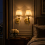 Two matching wall-mounted sconces with cream pleated drum shades and gold-toned bases, lit and mounted on a beige wall above a bedside table in a bedroom setting.