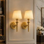 Two matching wall sconces with gold-toned ornate mounts and pleated cream fabric shades, mounted side by side on a decorative paneled wall in an interior setting.