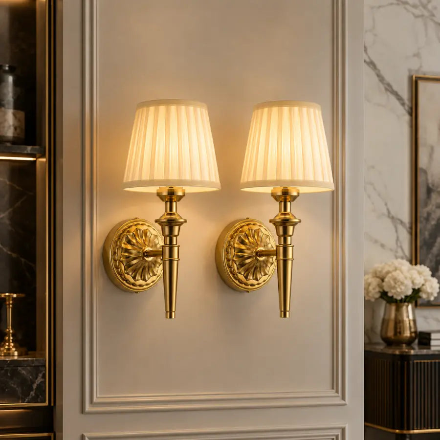Two matching wall sconces with gold-toned ornate mounts and pleated cream fabric shades, mounted side by side on a decorative paneled wall in an interior setting.