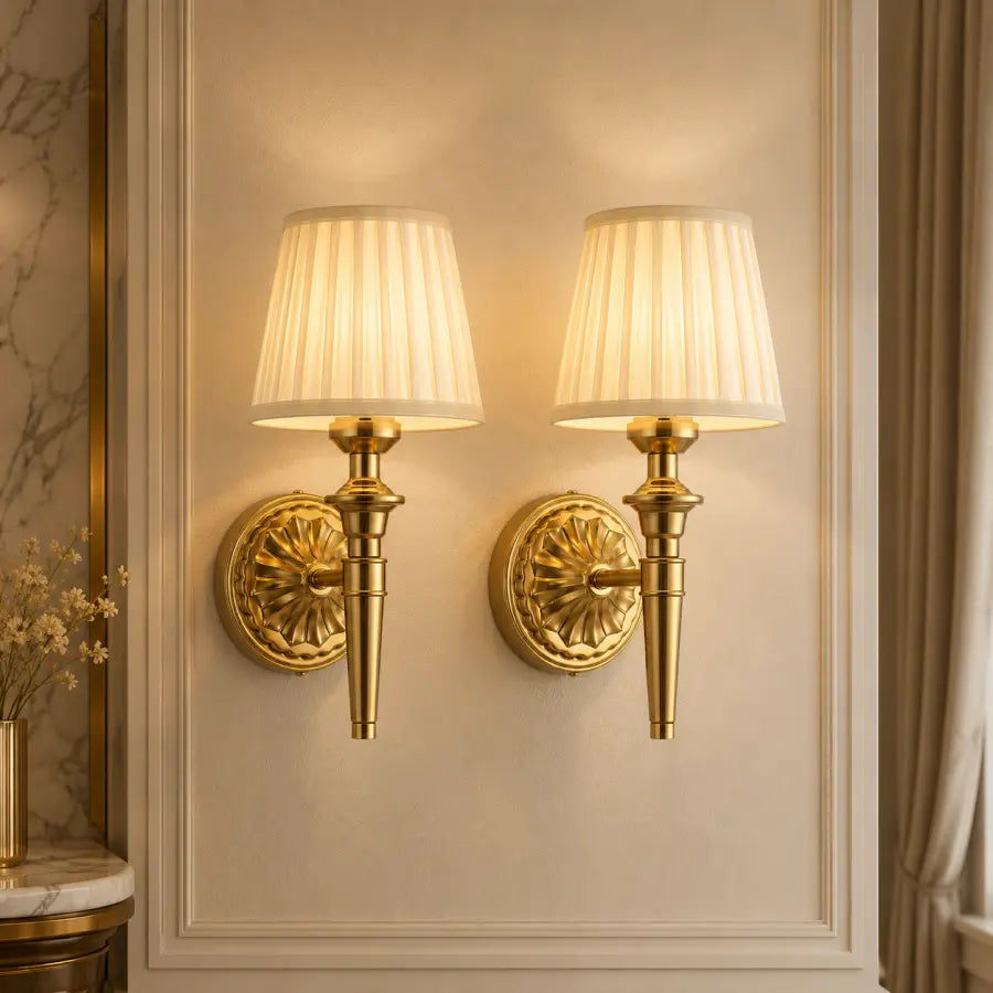 Two matching wall sconces with pleated cream fabric shades and ornate gold metal bases mounted on a beige paneled wall.