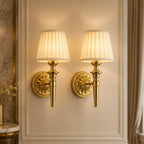 Two matching wall sconces with pleated cream fabric shades and ornate gold metal bases mounted on a beige paneled wall.