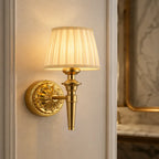 A classic brass wall sconce with a pleated cream fabric shade, mounted on a white paneled wall next to a marble-framed mirror.