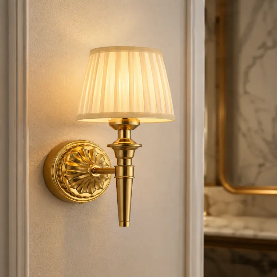 A classic brass wall sconce with a pleated cream fabric shade, mounted on a white paneled wall next to a marble-framed mirror.