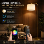 A smart floor lamp with a warm white drum shade and gold-toned metal stand in a living room setting, with hands holding a smartphone and a remote control below. Text on the left highlights phone and remote control features including brightness >1200 LM, Amazon Alexa compatibility, Google Assistant compatibility, and stepless dimming.