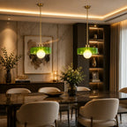 Two modern pendant lights with translucent green glass shades and gold-toned hardware hang over a dark marble dining table with cream upholstered chairs in a luxurious, warmly lit dining room.