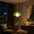 A modern pendant light with a translucent green glass shade and a gold-finished brass fixture hanging in a stylishly decorated living room with warm ambient lighting.