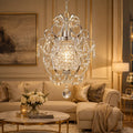 A large crystal chandelier with a silver-toned metal frame and hanging clear glass drops, suspended in a luxurious living room with a beige sofa, marble coffee table, and warm wall paneling.