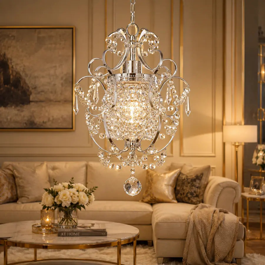 A large crystal chandelier with a silver-toned metal frame and hanging clear glass drops, suspended in a luxurious living room with a beige sofa, marble coffee table, and warm wall paneling.