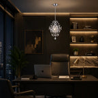 A luxury crystal chandelier with a silver-toned metal frame hangs over a modern home office desk setup. The room features dark wood paneling, a leather office chair, a laptop on the desk, and built-in shelves with warm lighting in the background.