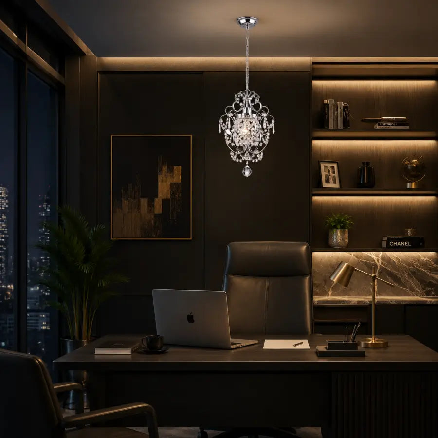 A luxury crystal chandelier with a silver-toned metal frame hangs over a modern home office desk setup. The room features dark wood paneling, a leather office chair, a laptop on the desk, and built-in shelves with warm lighting in the background.