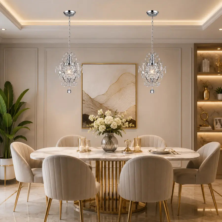 A modern dining room featuring two crystal chandeliers hanging above a round marble dining table with upholstered cream chairs. The room has warm beige walls, a framed abstract landscape print, and built-in shelving on the right.