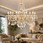 A large multi-arm gold and crystal chandelier hanging in a bright, elegant living room with a beige sofa, armchair, and decorative wall art.
