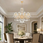 A large gold and crystal chandelier hangs over a dining table in a beautifully decorated elegant dining room with beige chairs, white flowers, and soft warm lighting.