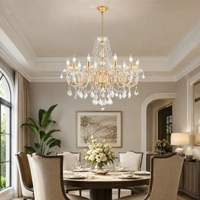 A large gold and crystal chandelier hangs over a dining table in a beautifully decorated elegant dining room with beige chairs, white flowers, and soft warm lighting.