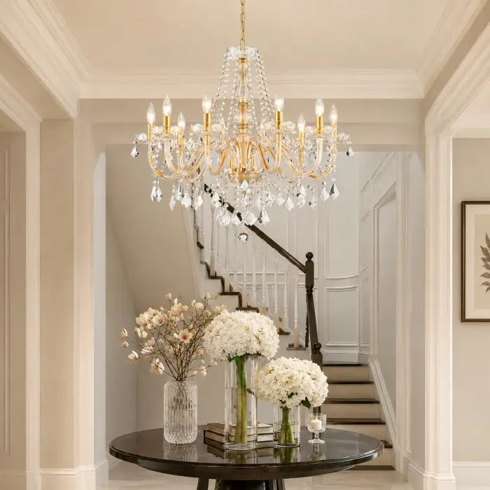 A large gold and crystal chandelier hangs in a bright, elegant hallway above a round black table with white flower arrangements, with a staircase visible in the background.