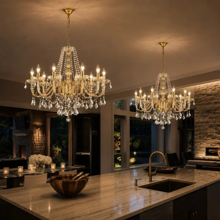 Two ornate gold and crystal chandeliers hanging in a modern kitchen with a marble island, stone wall, and warm ambient lighting.