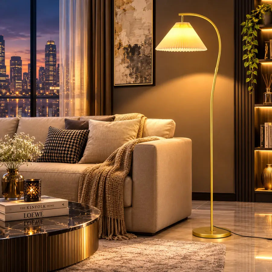 A modern floor lamp with a curved gold metal stand and a warm white shade is turned on in a stylishly decorated living room next to a beige sofa and a city skyline at dusk.