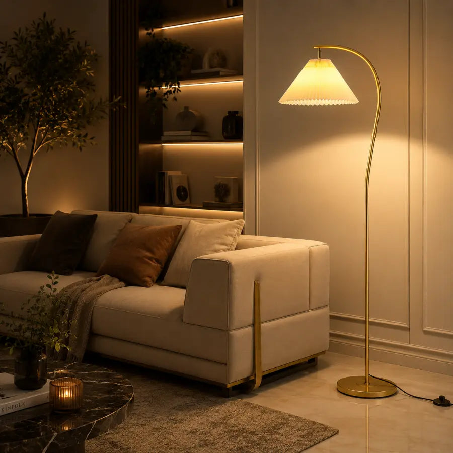 A modern floor lamp with a curved gold metal stand and a white pleated shade, glowing with warm light in a stylishly decorated living room next to a cream sofa.