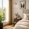 A modern floor lamp with a brushed gold metal frame and a clear cylindrical glass shade stands beside a bed in a softly lit bedroom. The room includes beige curtains, a wooden nightstand, a framed forest print on the wall, and a potted plant near the window.