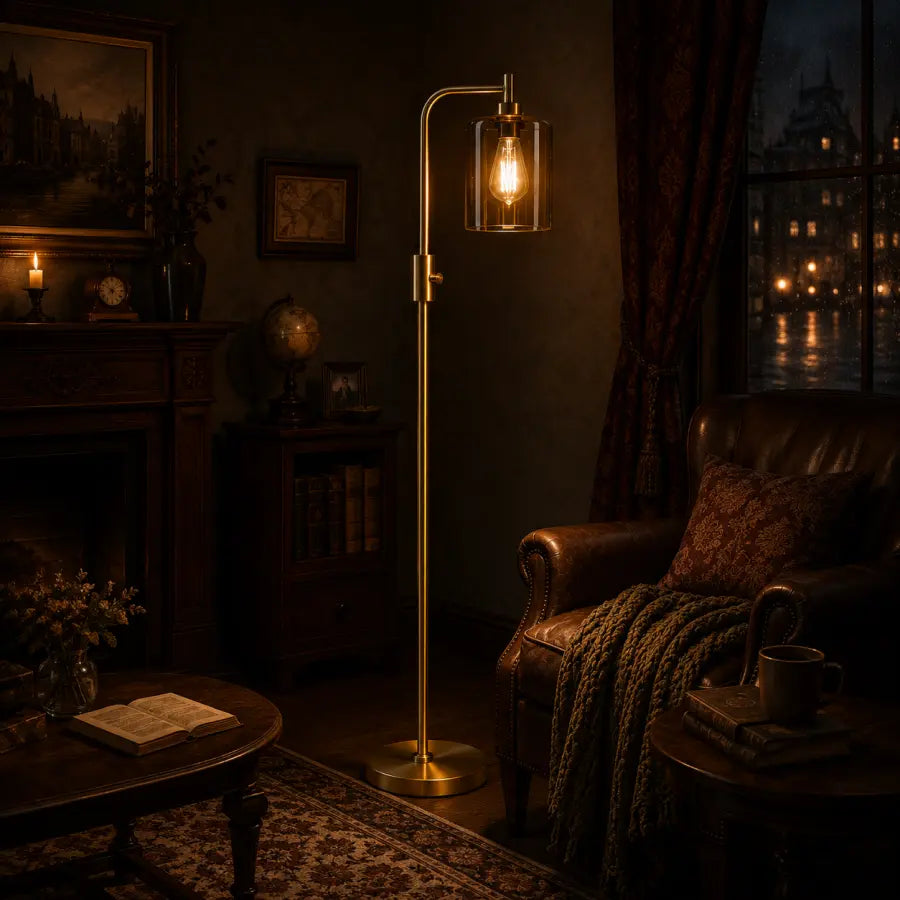A brass floor lamp with an arched arm and a clear cylindrical glass shade glowing with an Edison bulb, placed in a dark, cozy living room next to a leather armchair and window.