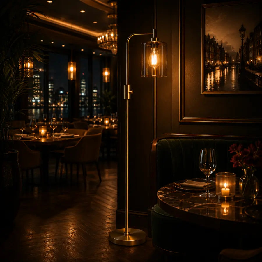 A modern floor lamp with a slim brass stand and a clear cylindrical glass shade housing a warm glowing Edison bulb, positioned in a dimly lit, elegant restaurant or lounge setting.