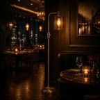A modern floor lamp with a slim brass stand and a clear cylindrical glass shade housing a warm glowing Edison bulb, positioned in a dimly lit, elegant restaurant or lounge setting.