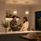Two modern pendant lights with gold-toned metal frames and clear glass shades hang over a kitchen island where a man and woman are standing and conversing in a warmly lit home interior.
