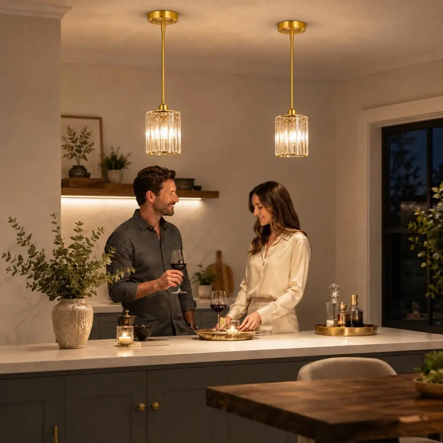 Two modern pendant lights with gold-toned metal frames and clear glass shades hang over a kitchen island where a man and woman are standing and conversing in a warmly lit home interior.