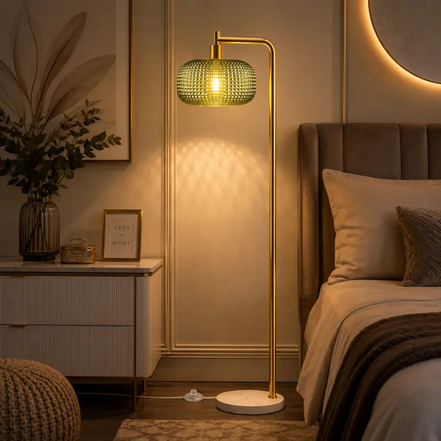 A modern floor lamp with a gold-finished metal frame and a textured green glass shade, glowing with warm light in a styled bedroom setting next to a bed.