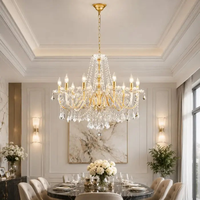 A large gold and crystal chandelier hangs over a dining table in an elegant, well-lit dining room with white walls, wall sconces, and a marble artwork.