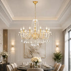 A large gold and crystal chandelier hangs over a dining table in an elegant, well-lit dining room with white walls, wall sconces, and a marble artwork.