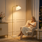 A woman in a cream-colored sleeveless dress sits in a beige armchair, looking at her phone in a warmly lit living room. A tall floor lamp with a white bell-shaped shade and gold frame stands next to her, casting a soft glow.