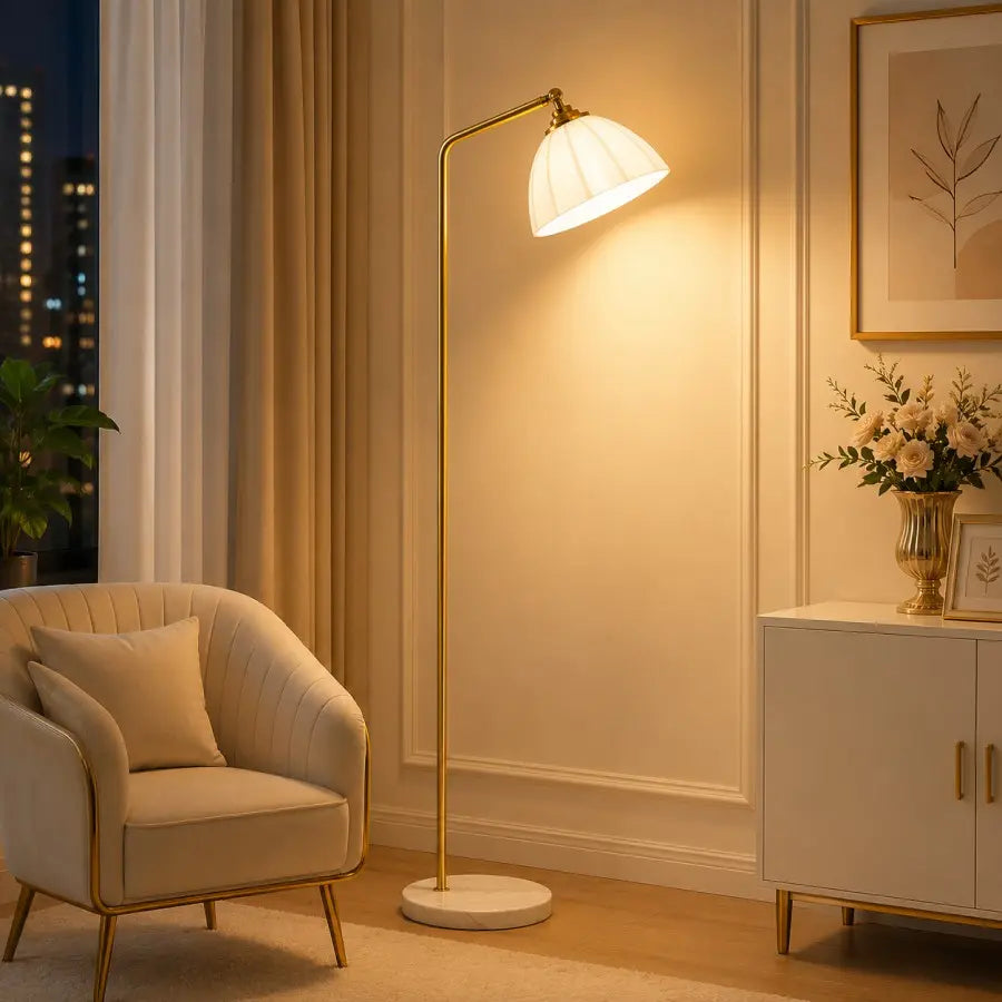 A modern floor lamp with a white pleated shade and a slim gold metal stand is lit in a warm, elegant living room setting next to a cream-colored armchair and a white sideboard.