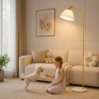 A warm, modern living room scene featuring a young girl kneeling on a beige carpet and playing with a small white poodle dog. To her right stands a tall floor lamp with a gold-finished metal frame and a white pleated shade.