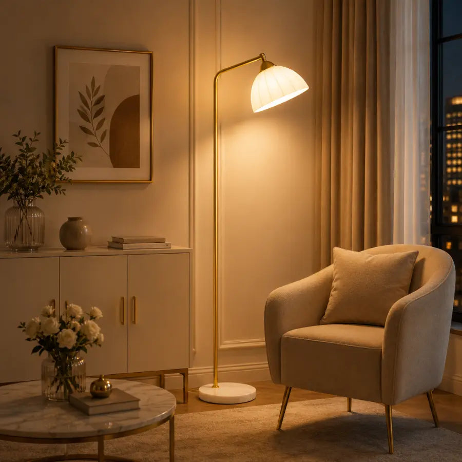 A modern floor lamp with a warm glowing white shade and gold-toned metal frame stands beside a beige armchair in a softly lit living room. The scene includes a cabinet, framed wall art, flowers, and a window with city lights at night.