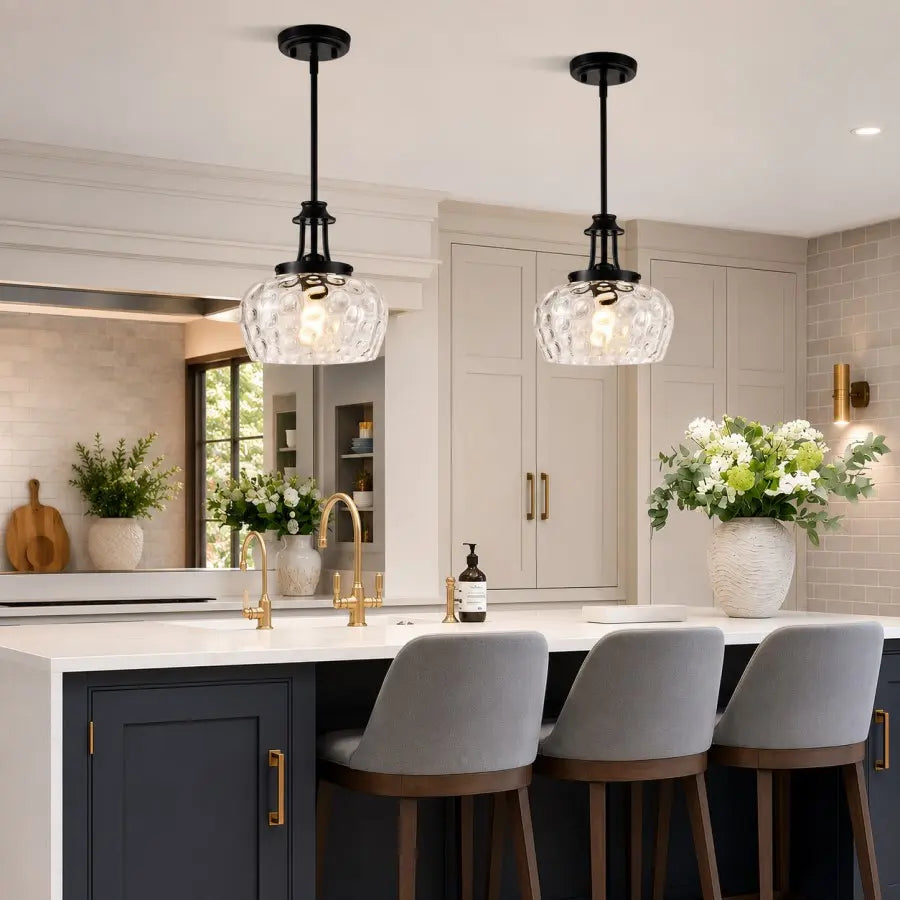 Two modern pendant lights with black metal stems and clear textured glass shades hanging over a kitchen island with gray bar stools.