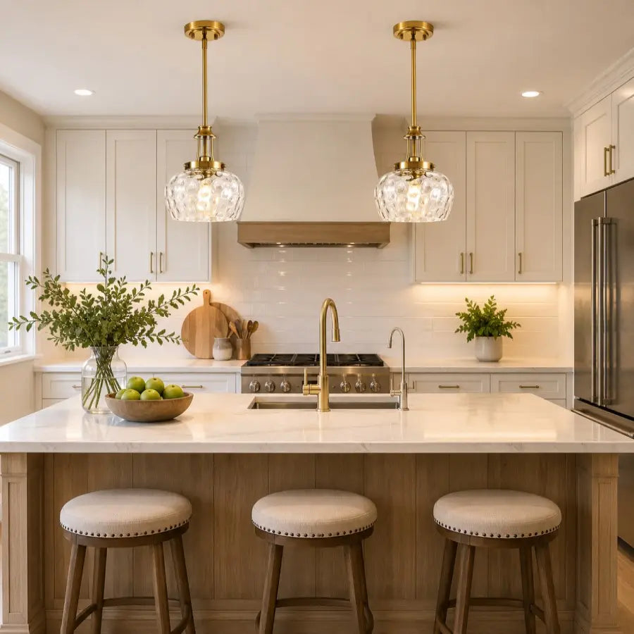 A modern kitchen island with two hanging pendant lights featuring clear glass globes and gold-toned metal frames. The scene includes white cabinetry, a marble countertop, a wood-look island base, three upholstered bar stools, a gas stove, and stainless steel appliances.