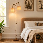 A rustic two-light floor lamp with a branch-like wooden stand and warm beige drum shades stands beside a bed in a softly lit bedroom. The room includes botanical wall art, white bedding, a patterned throw blanket, a plant, and a window.