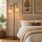 A rustic two-light floor lamp with a natural wood branch-style pole and beige fabric shades stands beside a neatly made bed in a softly lit bedroom. Framed botanical prints hang above the bed, and a potted plant sits near the window.
