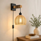 A wall-mounted pendant light with a black metal bracket, wooden accent, and a woven natural-fiber shade, shown lit in a warm indoor setting beside a table and vase of greenery.