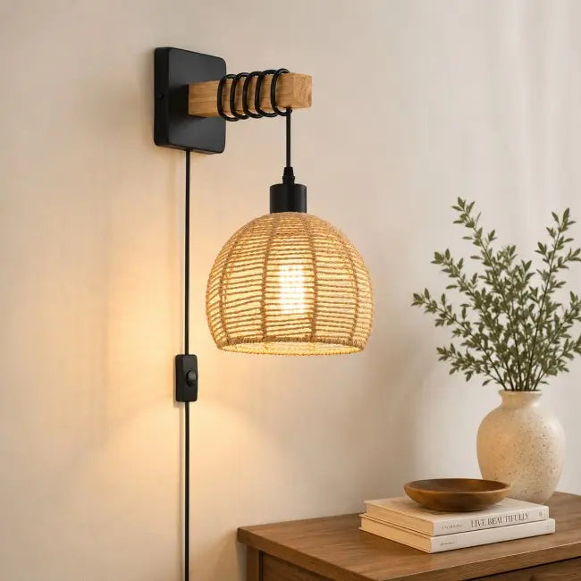 A wall-mounted pendant light with a black metal bracket, wooden accent, and a woven natural-fiber shade, shown lit in a warm indoor setting beside a table and vase of greenery.