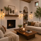 A cozy, warmly lit living room featuring a white stone fireplace with a dark wood mantel, a black fire pit, a wooden coffee table, and two cream-colored sofas with patterned throw pillows.