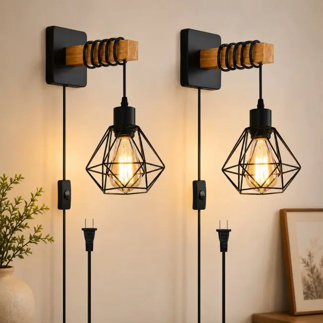 Two matching industrial-style wall sconces mounted on a beige wall, each with a black square backplate, wooden accent bar, coiled metal support, and a geometric black wire cage holding a glowing Edison bulb. Power cords and plugs are visible below, with a plant and framed picture in the room setting.