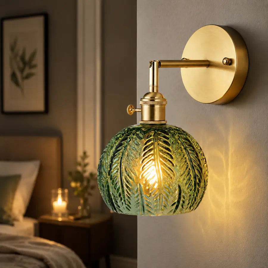 A wall-mounted sconce with a round green leaf-patterned glass shade and a brushed brass metal arm and backplate, shown lit in a bedroom setting.
